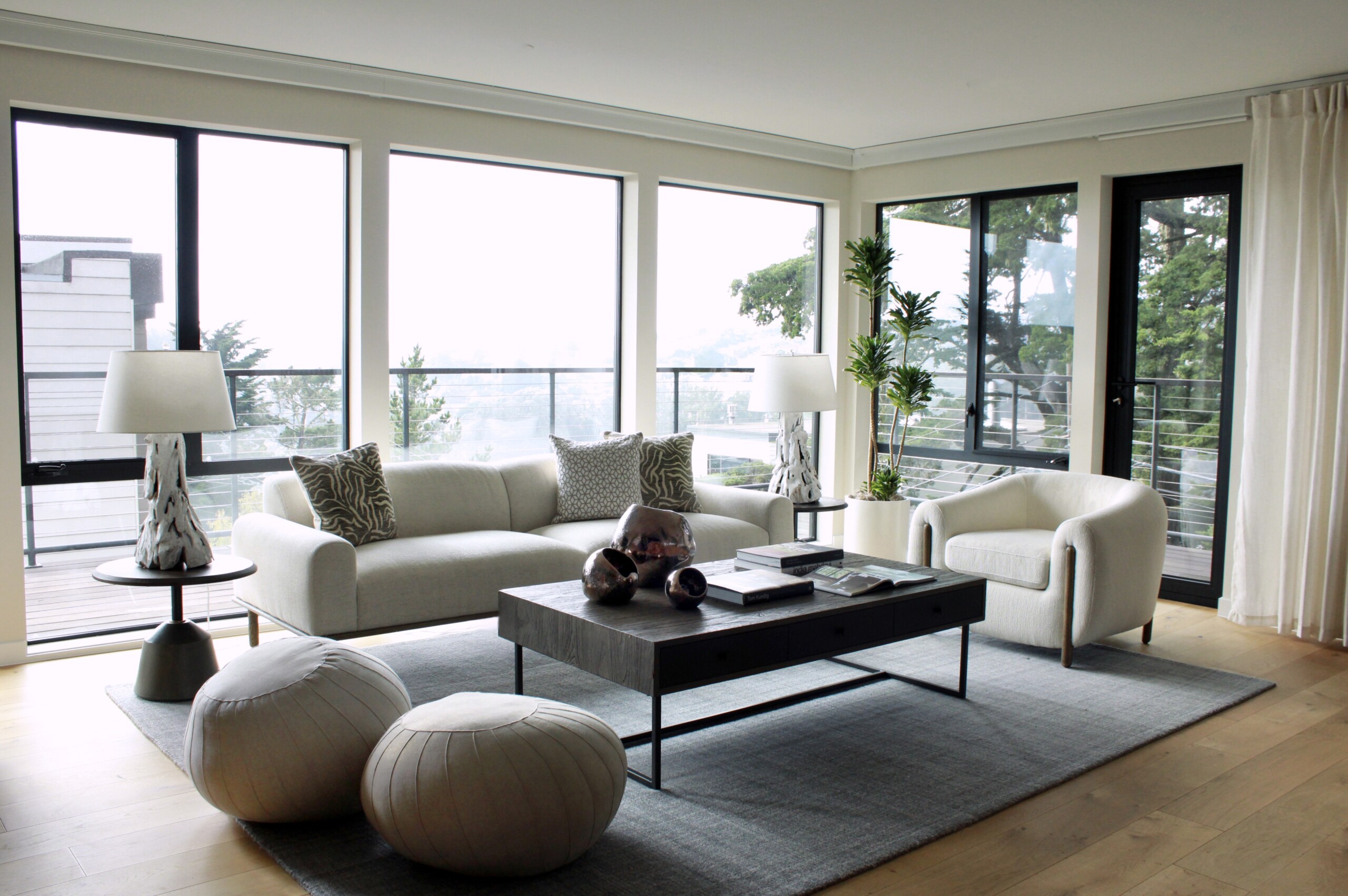 Modern Living Room With Large Windows, White Sofas, A Gray Rug, Round Poufs, A Black Coffee Table, And Potted Plants. Natural Light Fills The Space, Highlighting The Minimalistic Decor.