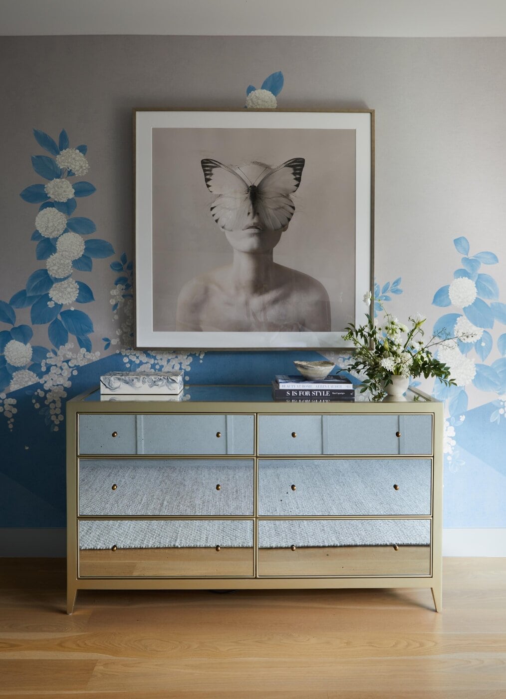 A Mirrored Dresser With Books, A Small Vase Of Flowers, And A Box On Top Stands Against A Wall With Blue Floral Wallpaper. Above The Dresser Hangs A Framed Black-And-White Portrait Of A Person With A Butterfly Over Their Face.