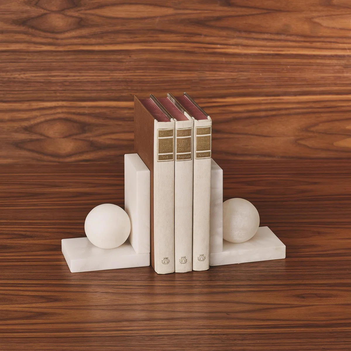 Three books with beige and brown covers are held upright by two white marble bookends, each featuring a rectangular base and a spherical accent, set against a wooden background.