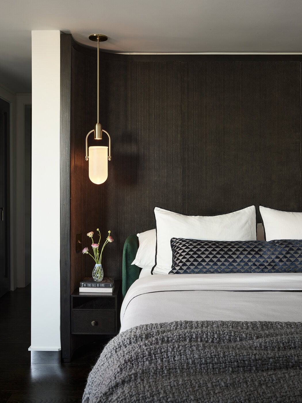 A Modern Bedroom Features A Bed With White Pillows, A Textured Gray Blanket, And A Patterned Bolster. A Glass Vase With Flowers And Books Sits On A Black Nightstand, Lit By A Gold Pendant Lamp Against A Dark Accent Wall.