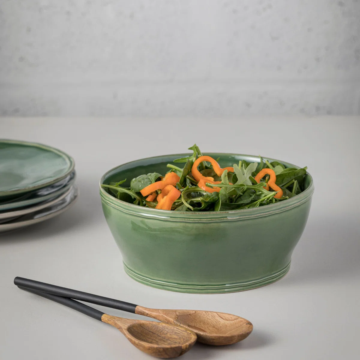 A green ceramic bowl filled with arugula and sliced yellow bell peppers sits on a light surface, next to a stack of green plates and two wooden serving utensils.
