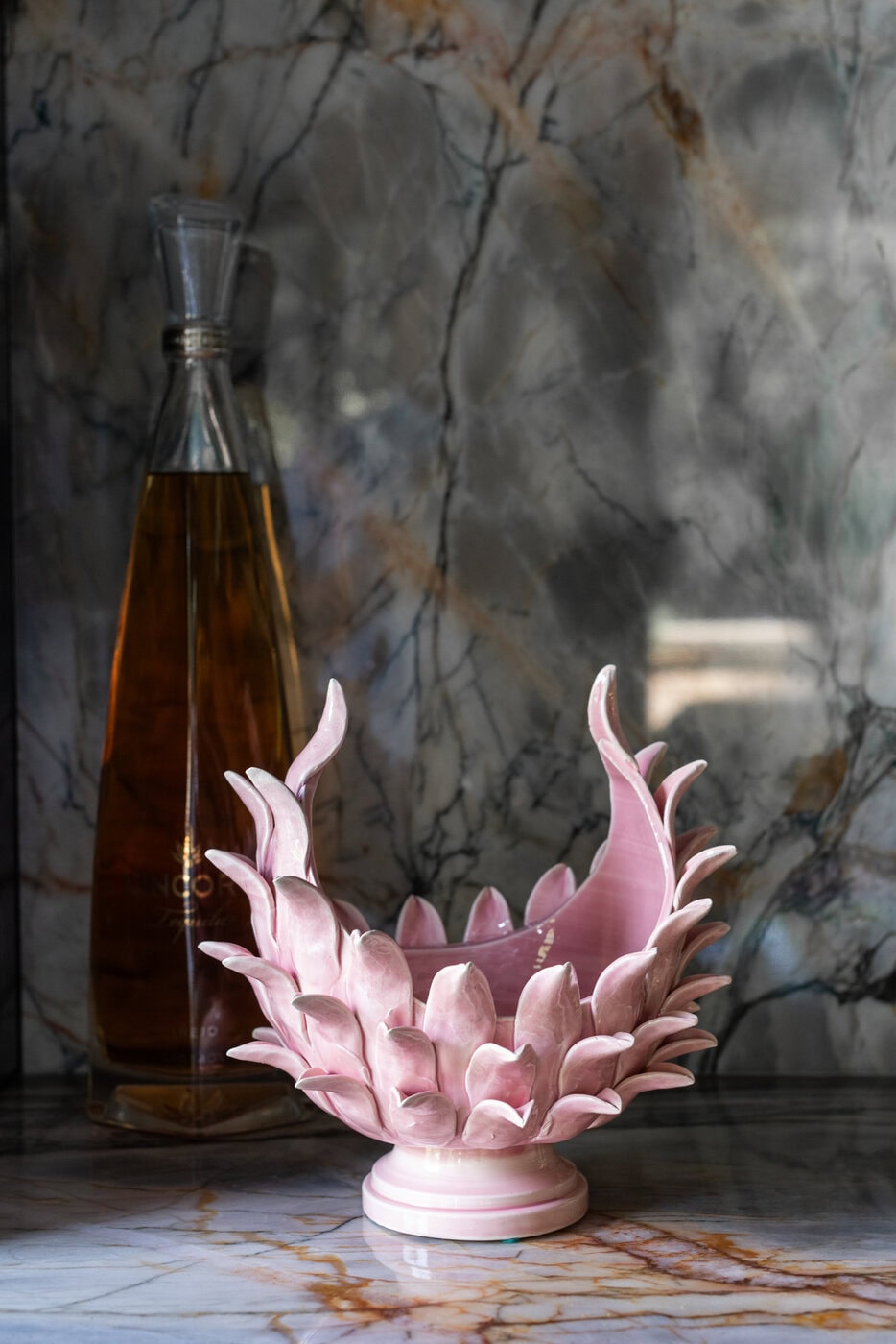 A Pink, Petal-Shaped Ceramic Bowl Sits On A Marble Surface In Front Of A Tall Glass Decanter Filled With Amber Liquid, Against A Gray And White Marbled Background.