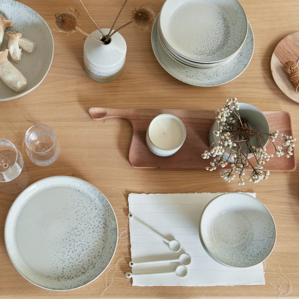 A wooden table set with speckled ceramic plates, bowls, a vase with dried flowers, a candle, glasses, measuring spoons, and decorative wooden and natural elements arranged neatly.
