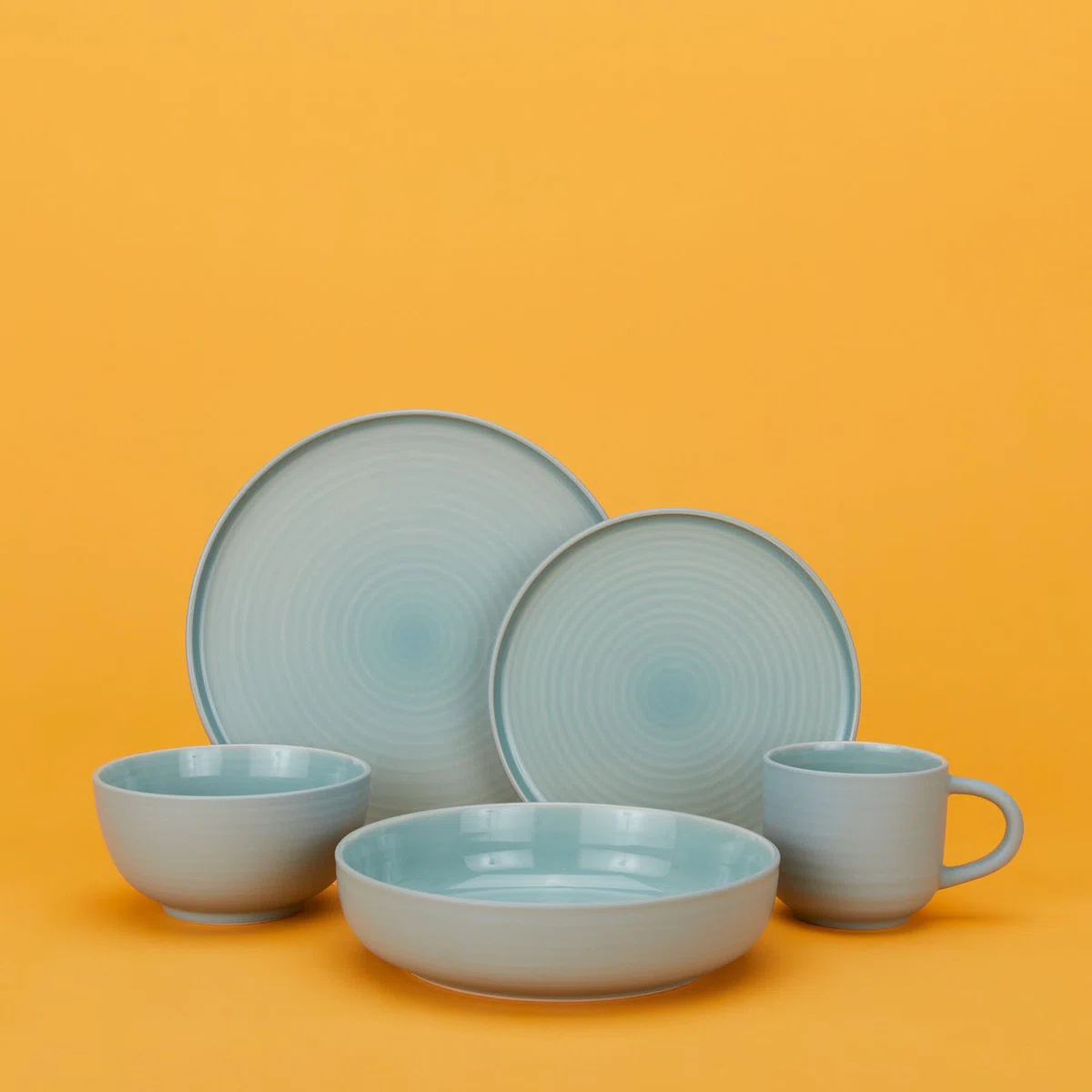 A set of light blue ceramic tableware, including two plates, a bowl, a mug, and a shallow dish, arranged against a solid orange background.