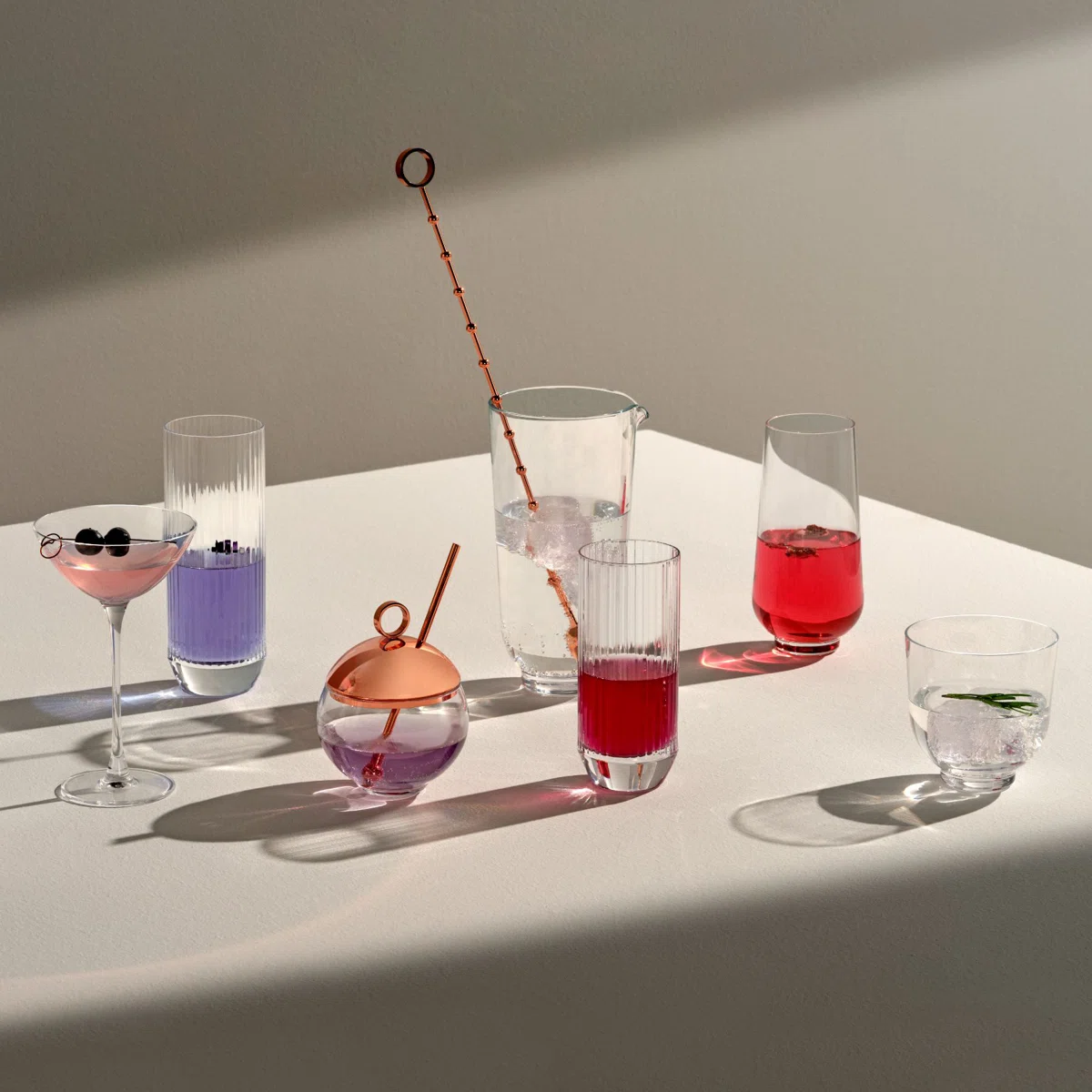 A variety of colorful drinks in different glassware, including a martini glass, highball glasses, and a spherical glass with a copper lid, are arranged on a white table in soft, natural light.