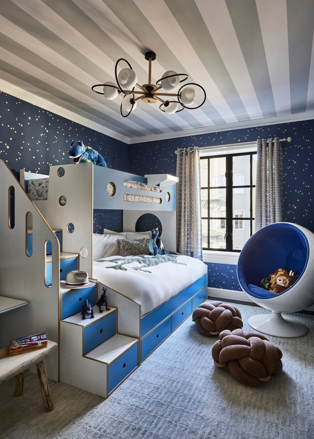 Children’s Bedroom With A Space Theme, Featuring A Blue And White Bunk Bed With Storage Stairs, Constellation-Patterned Wallpaper, A Modern Blue Egg Chair, Plush Knot Pillows, And Soft Natural Light From A Window.