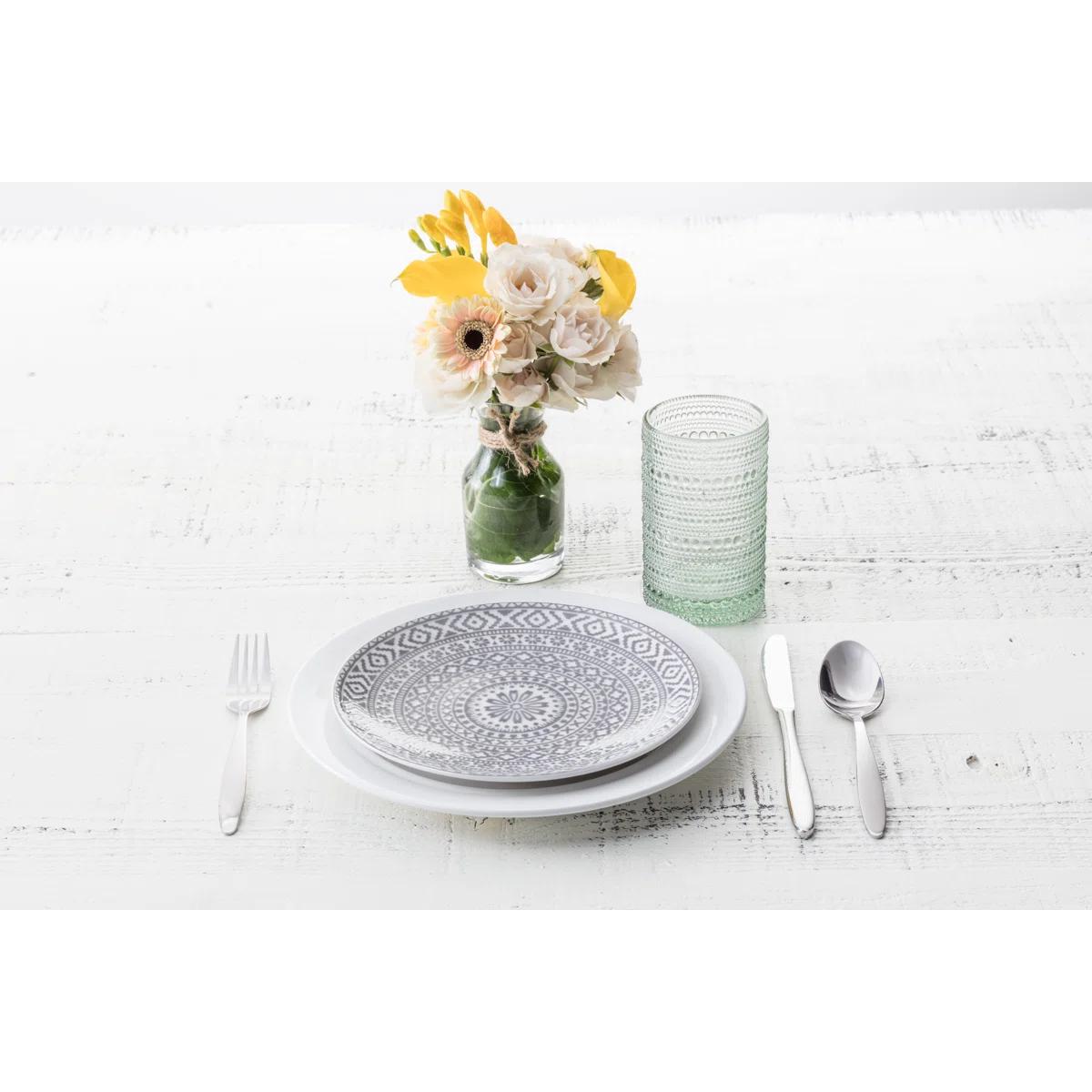 A table setting with a fork, knife, and spoon beside patterned plates, a textured green glass, and a vase of white and yellow flowers on a white wooden surface.