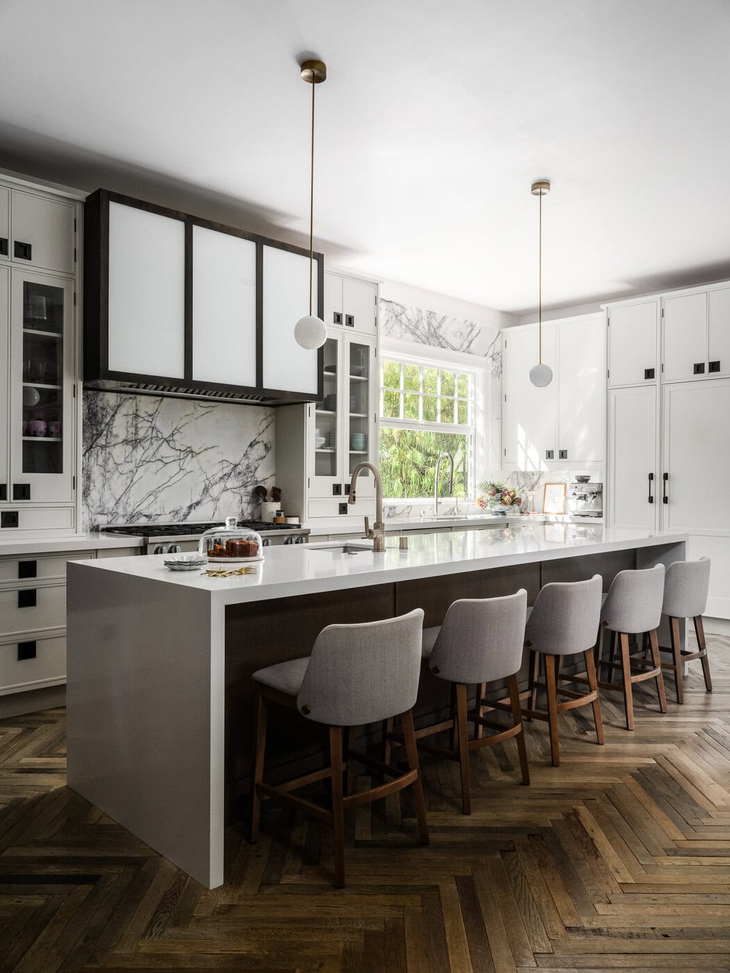 Modern Kitchen With A Large White Island, Five Upholstered Chairs, Marble Countertops And Backsplash, Pendant Lights, White Cabinets, A Window Above The Sink, And Herringbone Wood Floor.