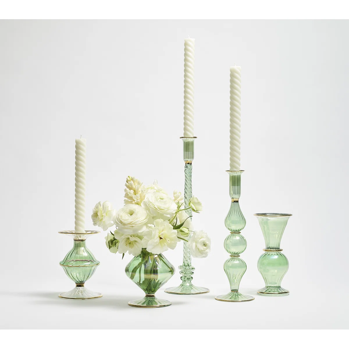 Five green glass candlestick holders, four with tall white candles and one holding a bouquet of white flowers, are arranged together against a plain white background.