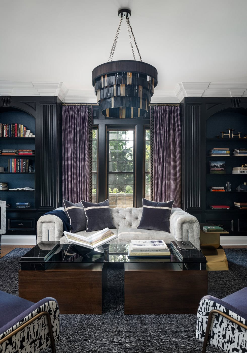 Elegant Living Room With A Tufted Gray Sofa, Dark Wood Coffee Tables, Navy Built-In Bookshelves, Purple Patterned Curtains, And A Modern Black And Gold Chandelier Centered Above. A Window Lets In Natural Light Behind The Sofa.