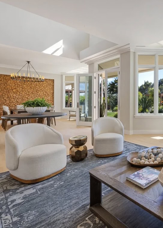 A Bright, Modern Living Room With Two White Swivel Chairs, A Wooden Coffee Table, And A Gray Rug. Large Windows And Open Doors Reveal A Lush Outdoor View. A Dining Area With A Log Accent Wall Is Visible In The Background.