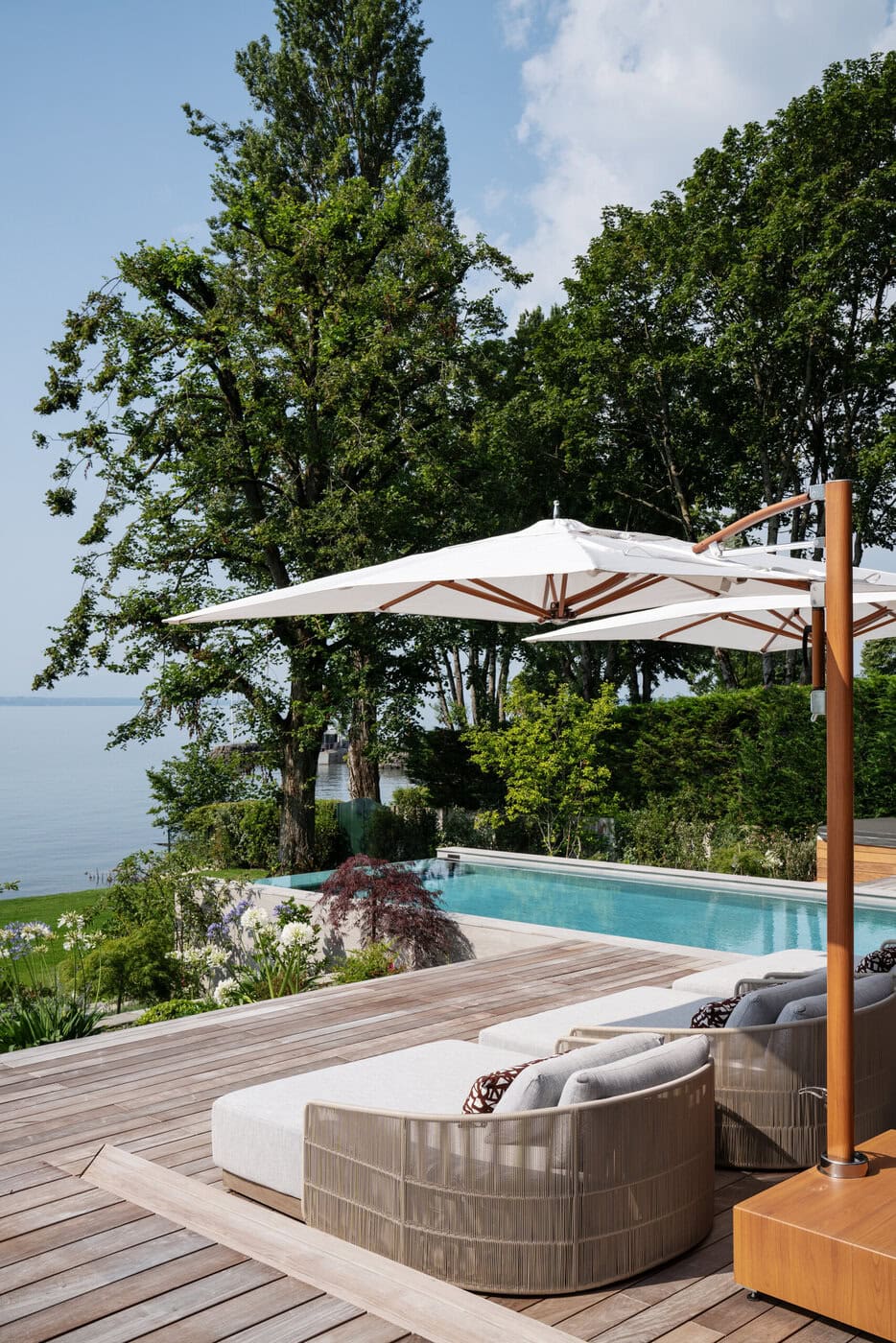 Modern Outdoor Patio With Lounge Chairs And A Large White Umbrella Beside An Infinity Pool, Overlooking A Scenic View Of Trees, Greenery, And A Body Of Water Under A Partly Cloudy Sky.