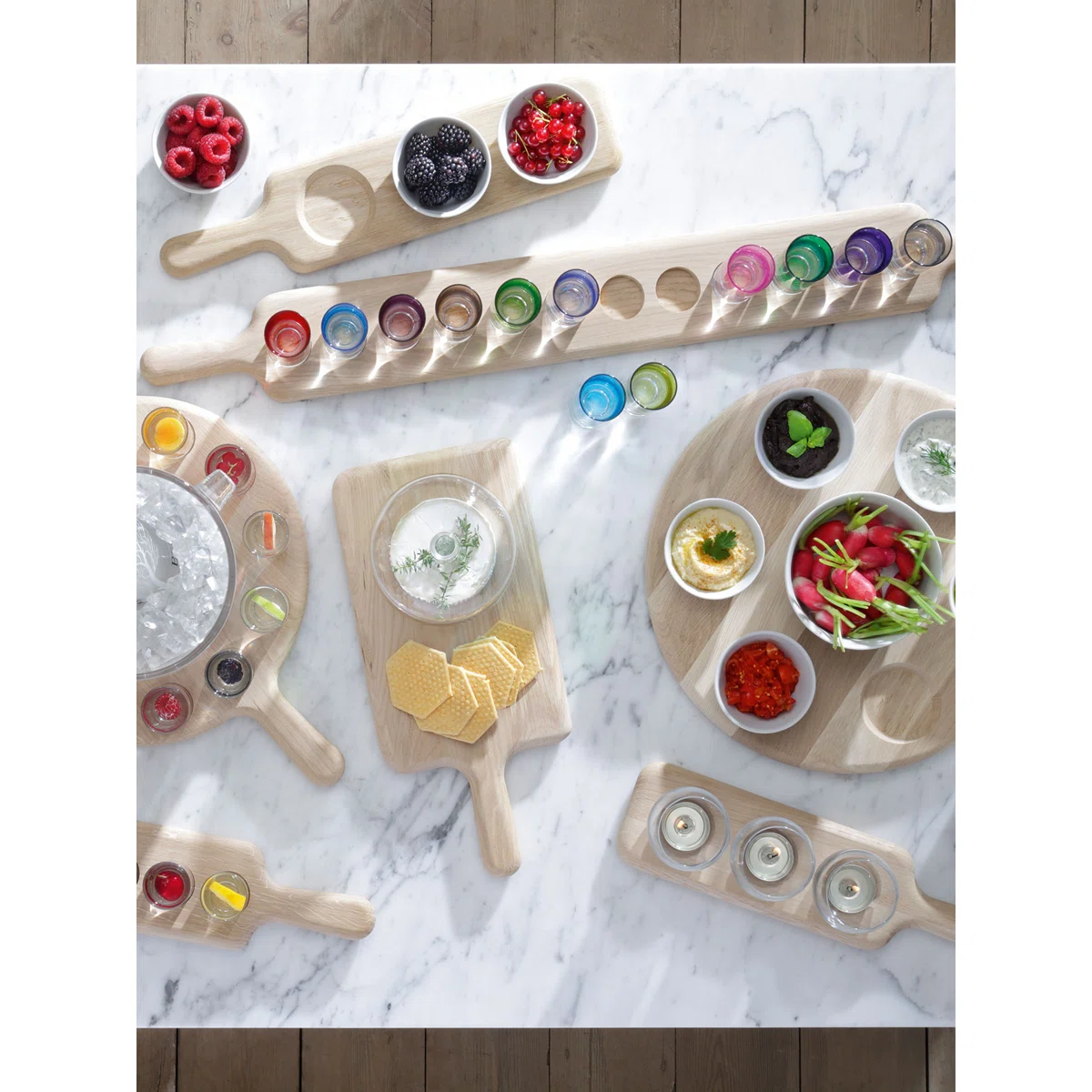 A top-down view of a marble table with wooden serving boards holding assorted colorful drinks, fresh vegetables, berries, cheese, and crackers, arranged for a stylish, casual gathering.