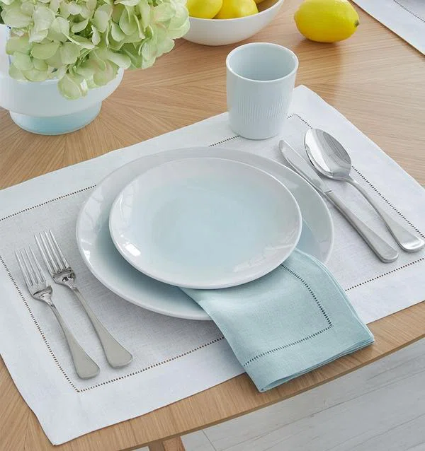 A table setting with white plates, silverware, a light blue napkin, a white mug, and a centerpiece of pale hydrangeas on a light wooden table with lemons in the background.