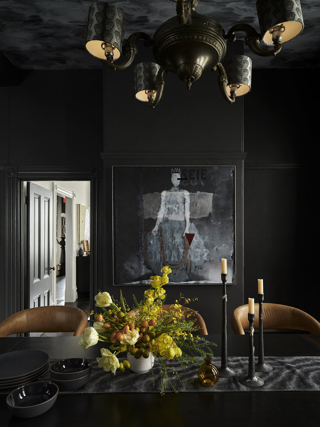 A Dark, Moody Dining Room With A Black Chandelier, Modern Art On The Wall, A Table Set With Black Dishes, Candles, And A Floral Arrangement Of Yellow And White Flowers. Tan Leather Chairs Surround The Table.
