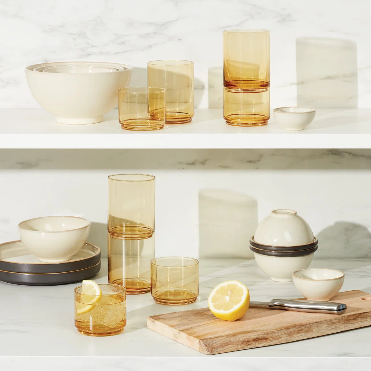 A kitchen scene with amber glasses, cream-colored bowls and plates, a wooden cutting board with a sliced lemon and knife, all arranged on white shelves against a marble background.