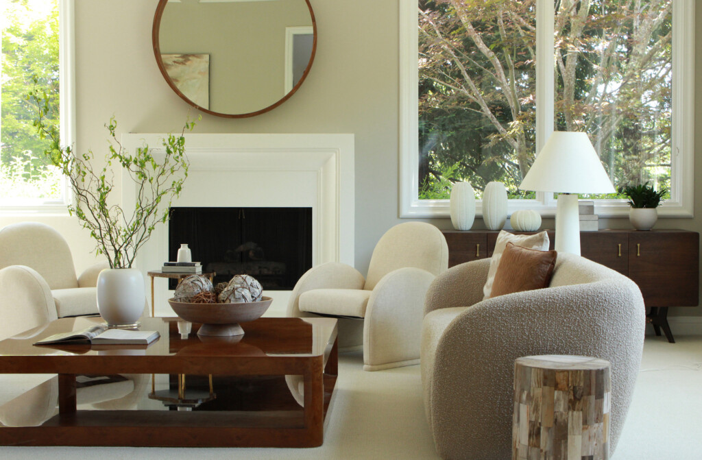 A modern living room with cream-colored curved chairs, a wooden coffee table, neutral decor, a round mirror above a white fireplace, large windows, and bright natural light.
