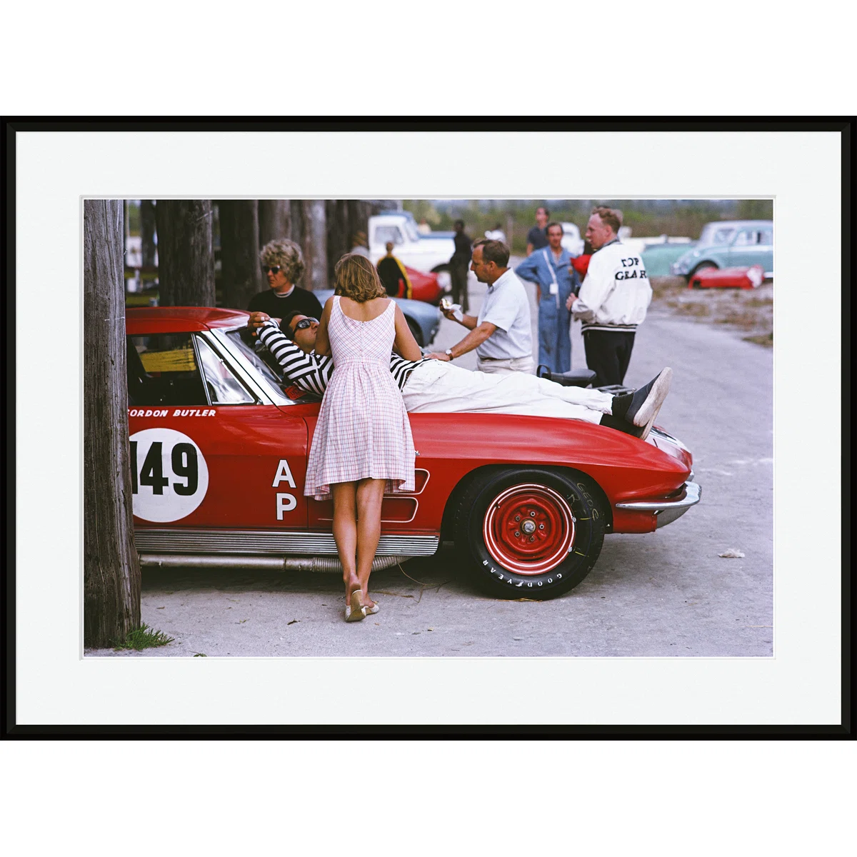 Several people gather around a vintage red race car numbered 149. A woman in a pink dress leans on the car, while others chat nearby. Trees and cars are in the background on a sunny day.