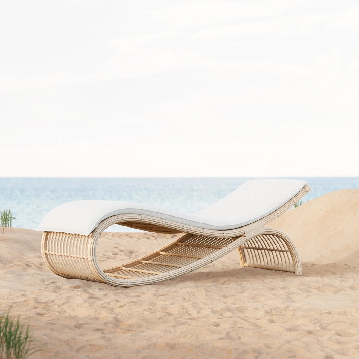 A curved, modern rattan lounge chair with a white cushion sits on sandy beach dunes, facing a calm blue sea under a bright sky.
