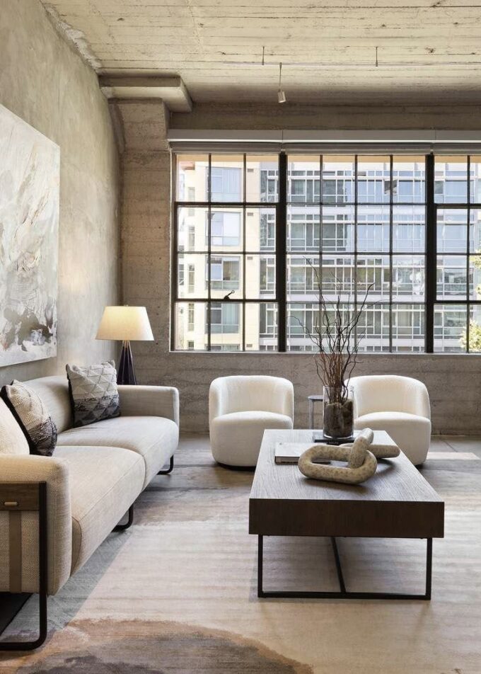 Modern living room with large industrial windows, neutral-toned furniture, a beige sofa, two white armchairs, a wooden coffee table with decorative objects, a floor lamp, and abstract wall art. Natural light fills the space.