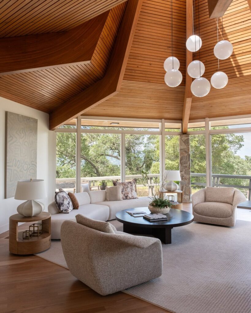 A Modern Living Room With Large Windows, Wood-Paneled Ceilings, A Round Black Coffee Table, A White Sofa, Two Beige Armchairs, And Contemporary Pendant Lights. Lush Greenery Is Visible Outside.