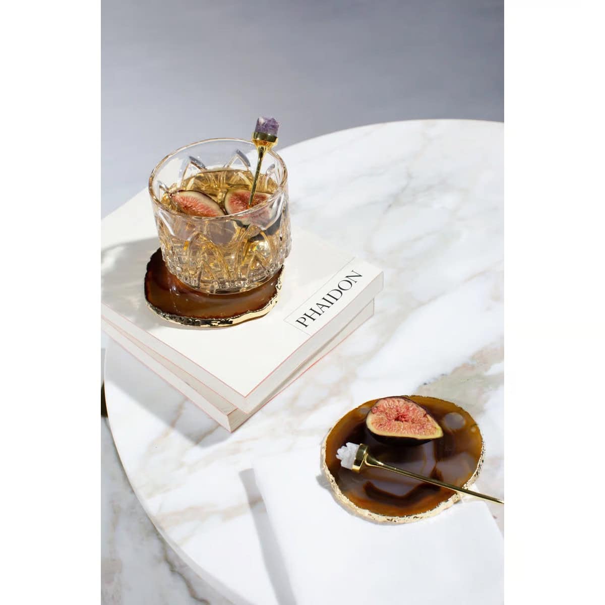 A crystal glass with a fig garnish sits on a book atop a marble table, alongside a drink coaster with a fig slice and gold spoon. Another coaster with a fig and spoon is on the table below.