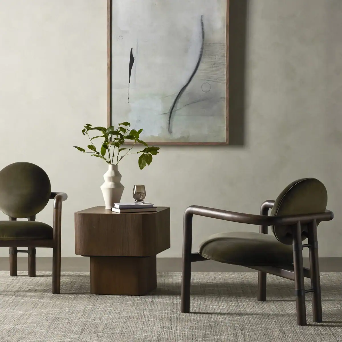 Two modern green velvet chairs with rounded backs and wood frames sit beside a dark wood side table with a white vase of greenery and a glass. Abstract art hangs on a neutral wall behind them.