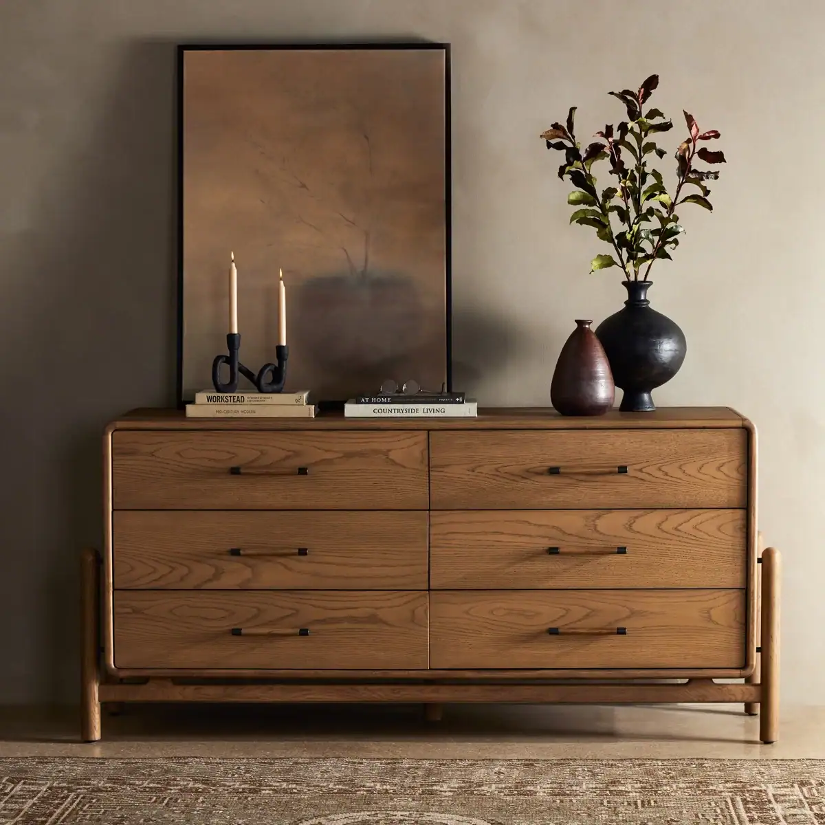 A wooden dresser with six drawers is topped with a framed artwork, two lit candles in dark holders, stacked books, and two vases—one brown and one black with leafy branches. The scene is set against a neutral wall.