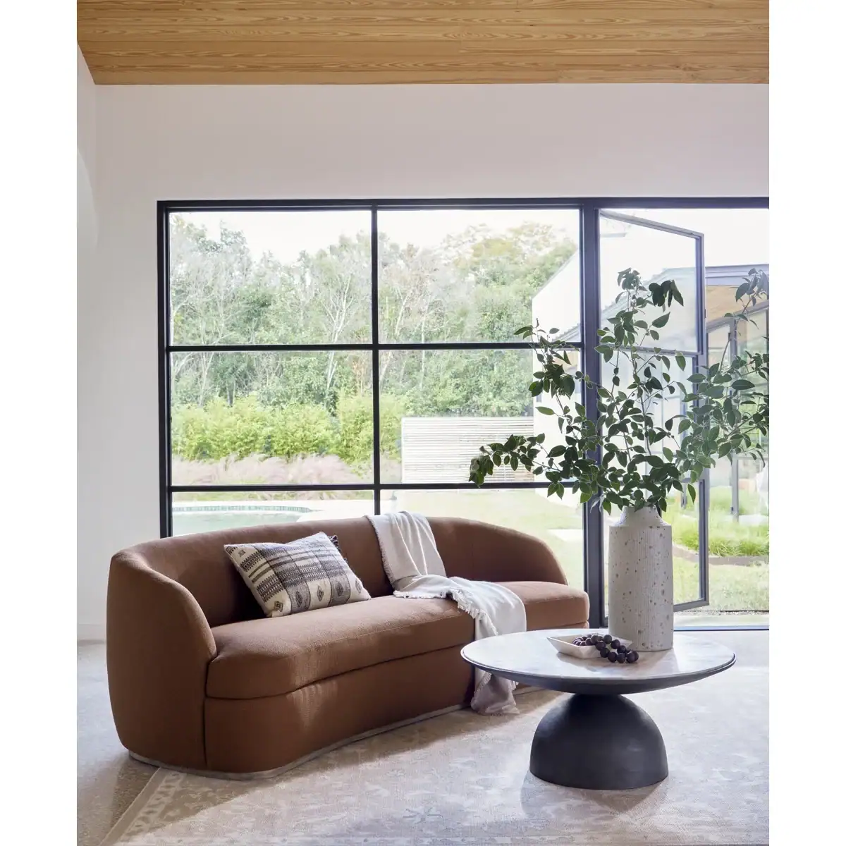 A modern living room with a curved brown sofa, patterned cushions, a round coffee table with a vase of leafy branches, and large windows overlooking greenery outside. Natural light fills the space.
