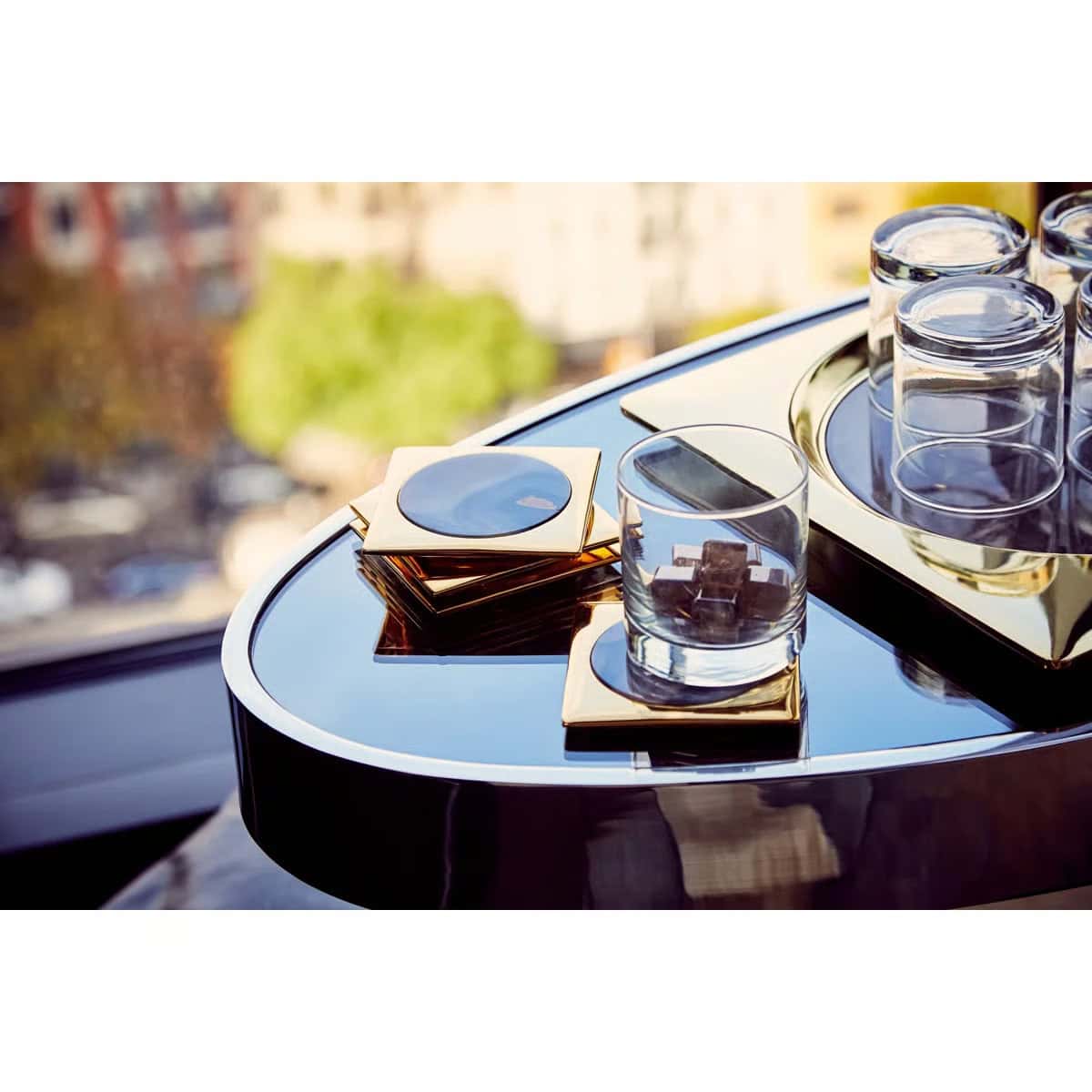 A close-up of a glossy black table with gold coasters, a glass with brown sugar cubes, and several stacked empty glasses, set near a window with a blurred cityscape in the background.