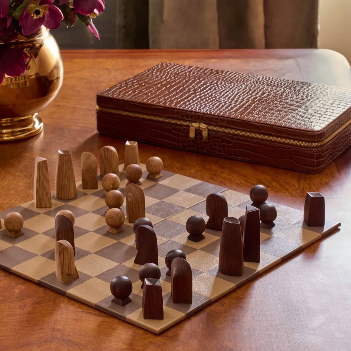 A modern chess set with uniquely shaped wooden pieces sits on a brown chessboard atop a wooden table. A textured brown case and a gold vase with flowers are in the background.