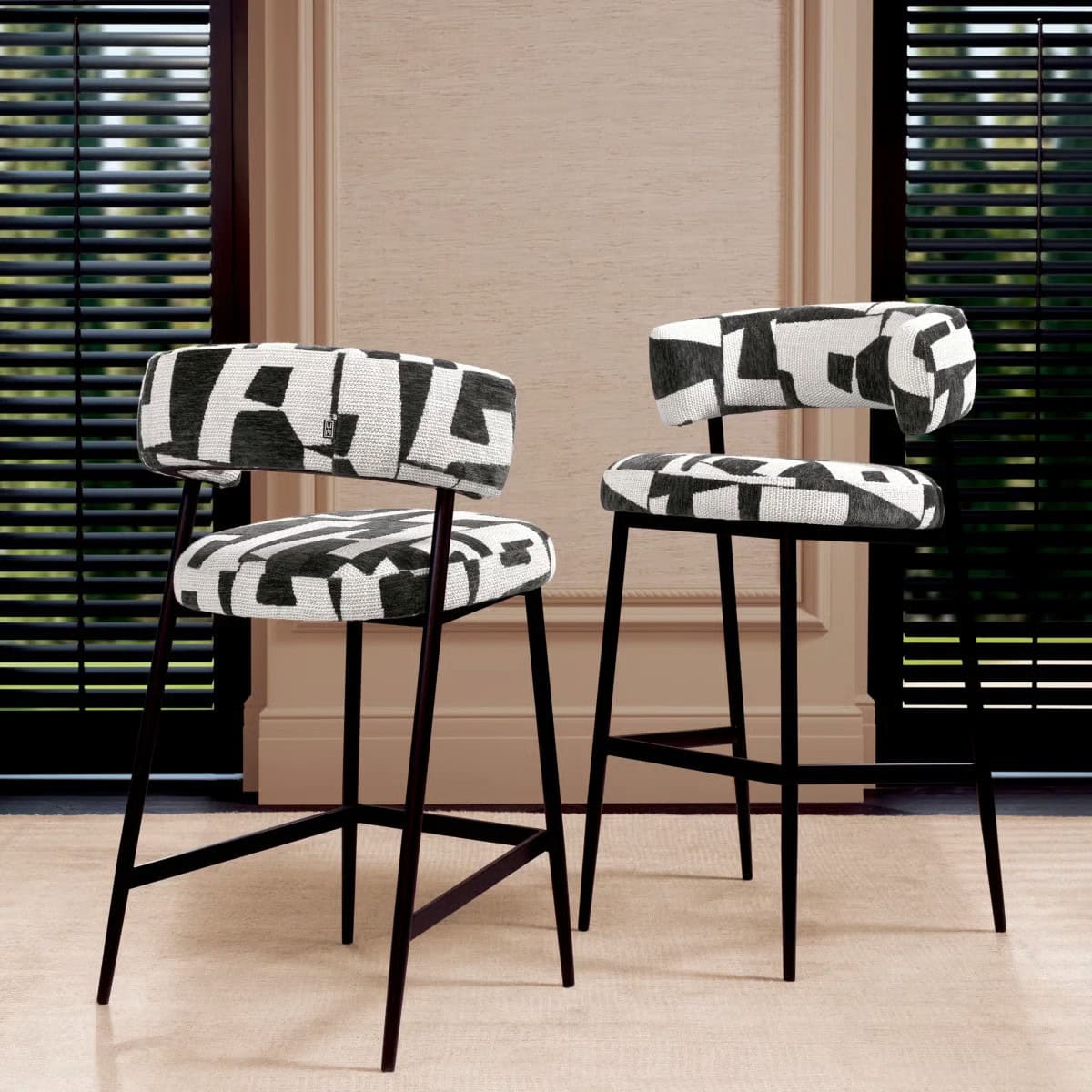 Two modern bar stools with black and white abstract patterned cushions and black metal legs are positioned on a beige carpet in front of tall windows with black blinds.