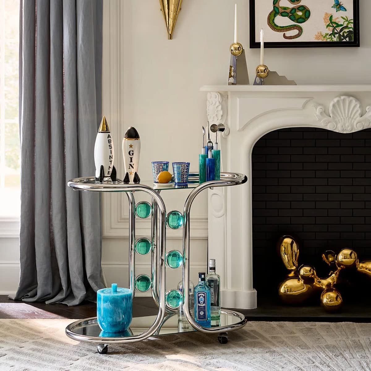 A modern bar cart with blue and silver accents holds bottles, glasses, and a blue candle in front of a white fireplace with a gold balloon dog sculpture and decorative objects on the mantel.