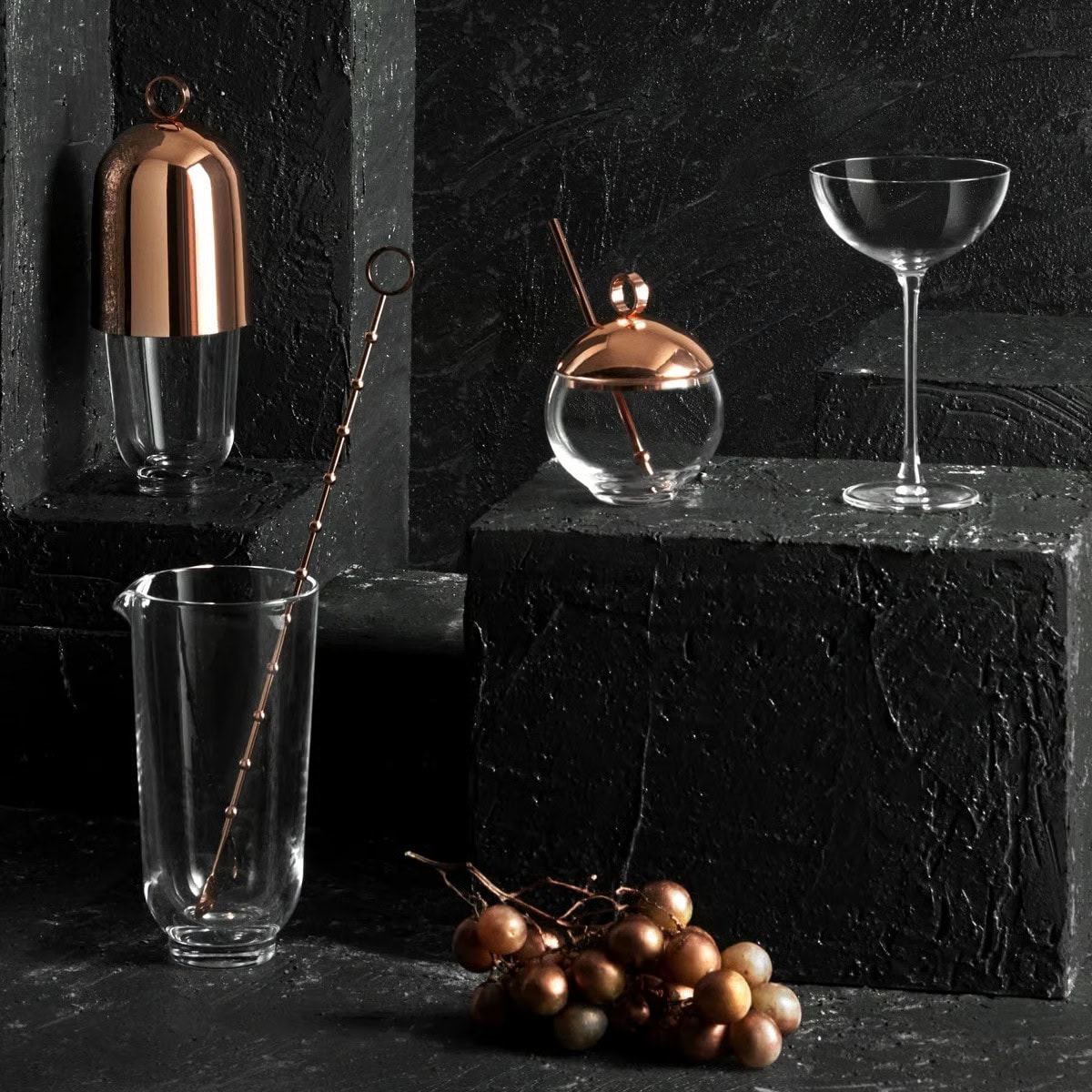 A stylish barware set on black stone blocks features a gold-accented shaker, a glass decanter with a gold lid and stirrer, a coupe glass, and a bunch of golden grapes in the foreground.
