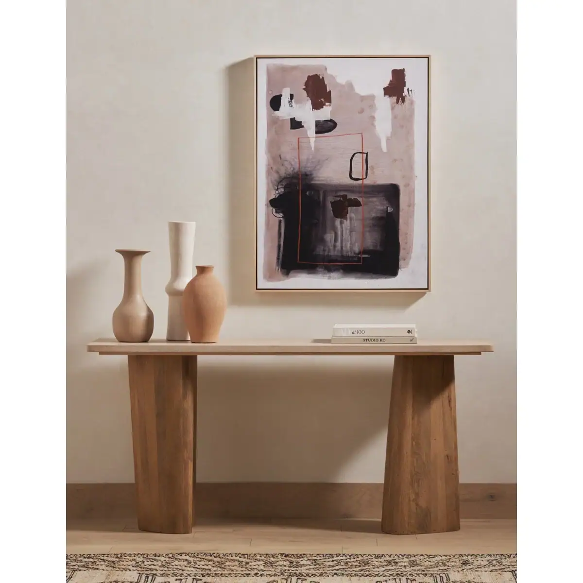 A wooden console table holds three ceramic vases and a closed book, with an abstract painting featuring brown, black, and white shapes hanging on the neutral-colored wall behind it.