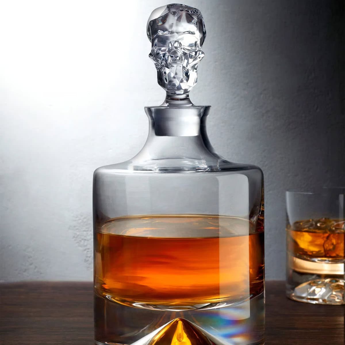 A crystal decanter filled with amber whiskey sits on a wooden surface, featuring an intricately sculpted stopper. In the background, a glass with whiskey and ice is partially visible.