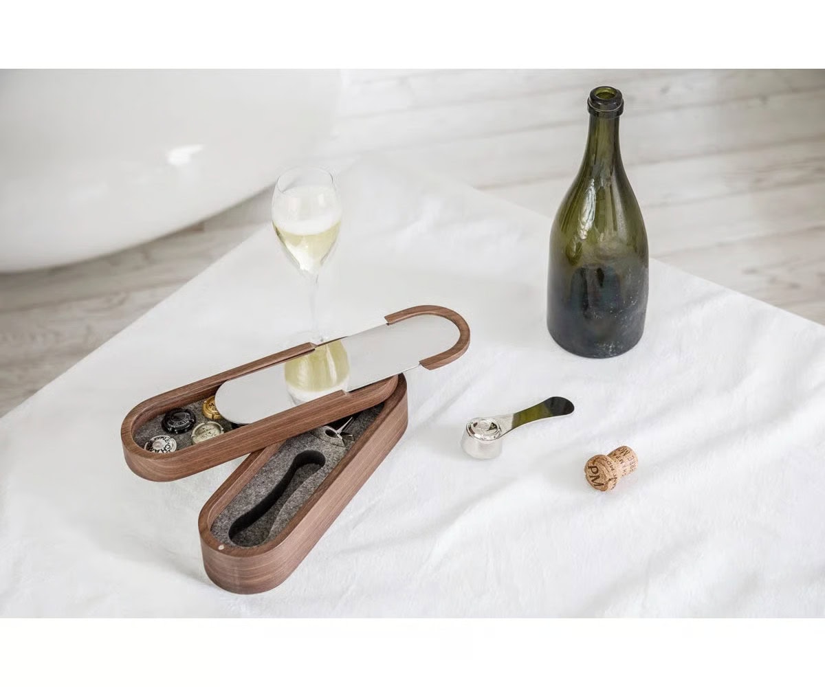 A wooden wine tool set with a mirrored lid lies open on a white tablecloth next to a glass of white wine, an empty wine bottle, a corkscrew, and a cork.