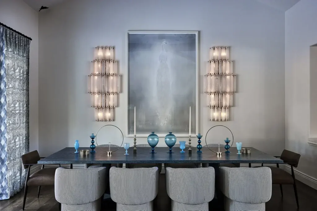 A Modern Dining Room With A Dark Table, Eight Light Upholstered Chairs, Blue Decorative Vases And Candles, Two Wall Sconces, And A Large Misty Artwork On A White Wall. Patterned Curtains Hang To The Left.