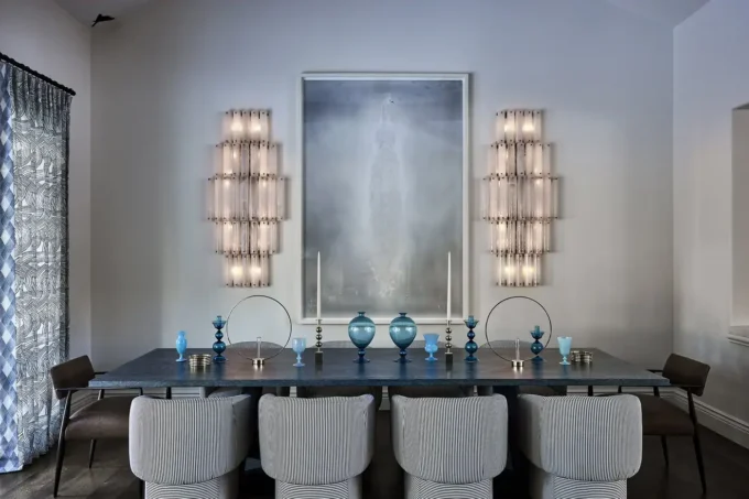A modern dining room with a dark table, eight light upholstered chairs, blue decorative vases and candles, two wall sconces, and a large misty artwork on a white wall. Patterned curtains hang to the left.