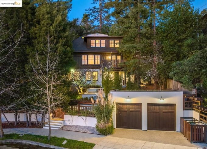 A large three-story house with many windows is surrounded by tall trees and greenery. In front, there is a white detached garage with two wooden doors, and a curved driveway leads up to the property at dusk.