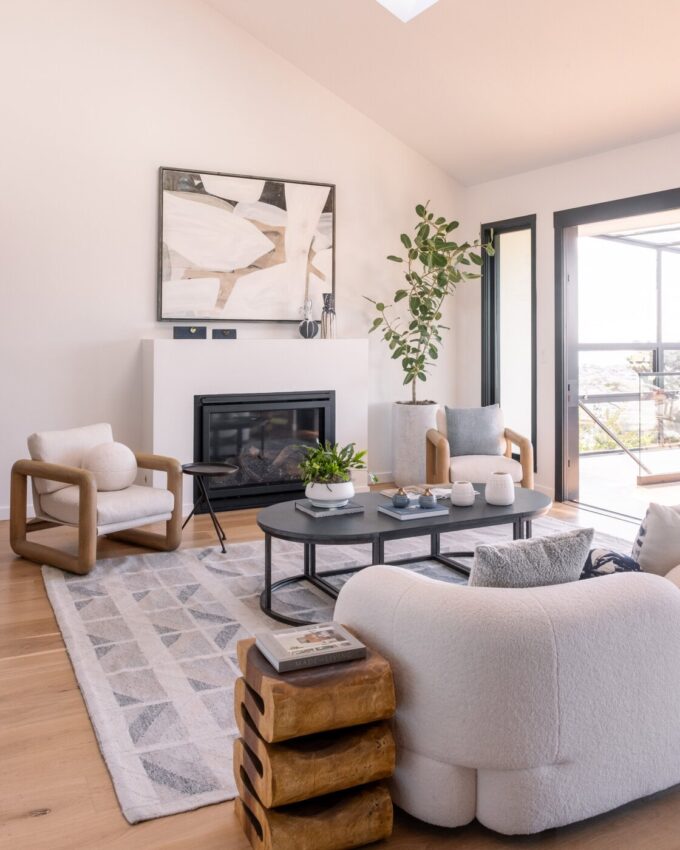 Modern living room with light wood floors, cozy white and beige armchairs, a round black coffee table, a patterned rug, and a fireplace with abstract art above it. Large windows bring in natural light and greenery.