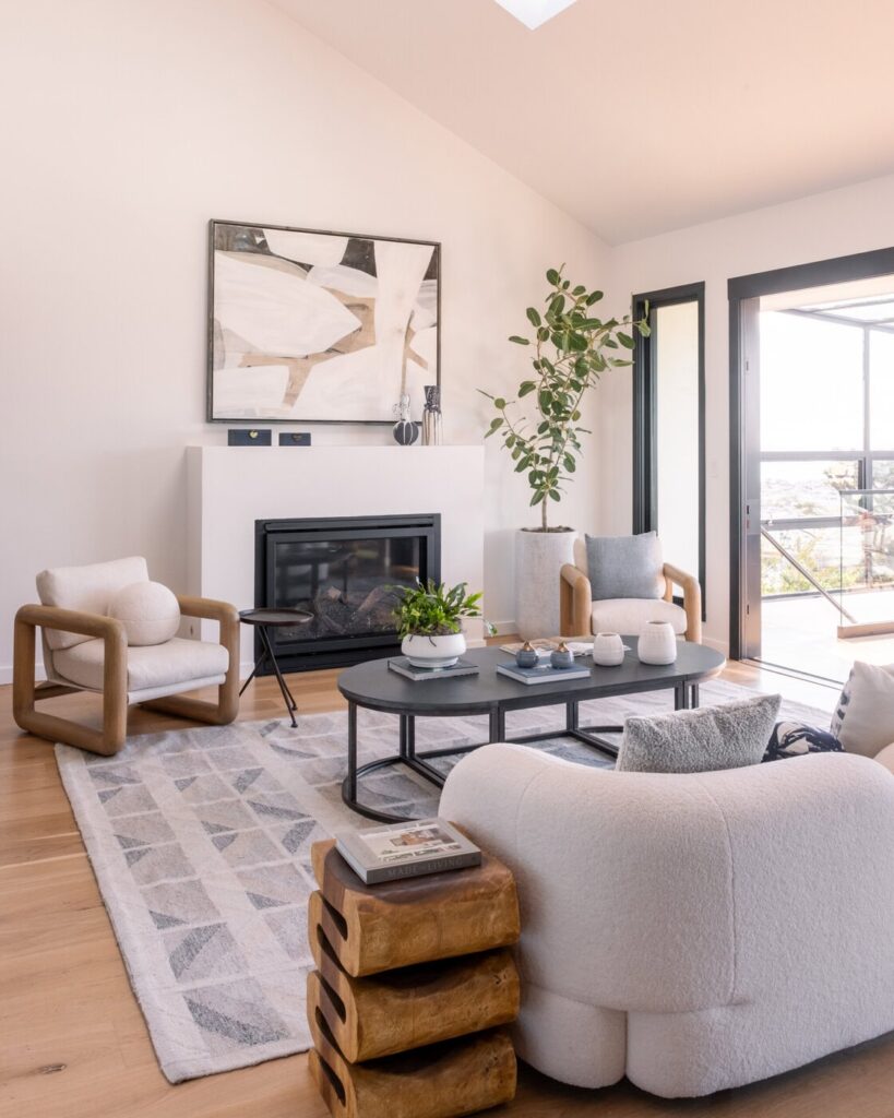 Modern Living Room With Light Wood Floors, Cozy White And Beige Armchairs, A Round Black Coffee Table, A Patterned Rug, And A Fireplace With Abstract Art Above It. Large Windows Bring In Natural Light And Greenery.
