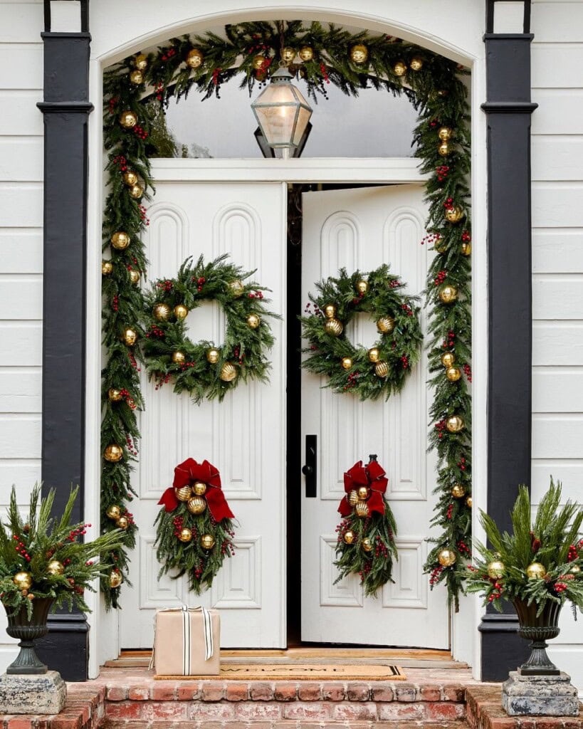 White Double Doors Decorated With Christmas Wreaths And Garlands Featuring Red Bows, Gold Ornaments, And Greenery; Two Planters With Seasonal Foliage And A Single Wrapped Gift Sit On The Brick Doorstep.