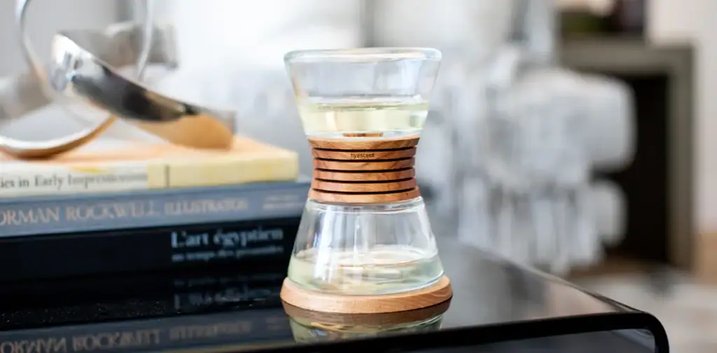 A Clear Hourglass-Shaped Glass With A Wooden Ring And Base Sits On A Black Table, Next To Stacked Books And Modern Decor In A Cozy, Softly Lit Room.