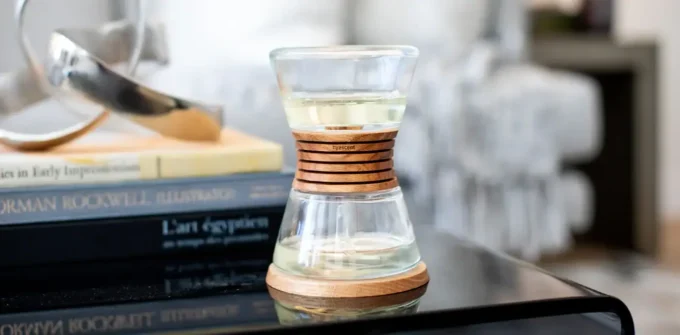 A clear hourglass-shaped glass with a wooden ring and base sits on a black table, next to stacked books and modern decor in a cozy, softly lit room.