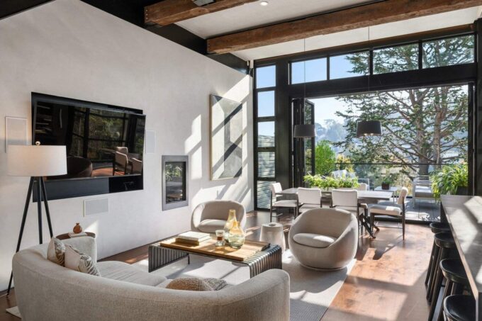 A modern living room with neutral tones, large windows, exposed beams, cozy seating, and a view of trees and mountains. Natural light fills the space, and a dining area is visible near the windows.