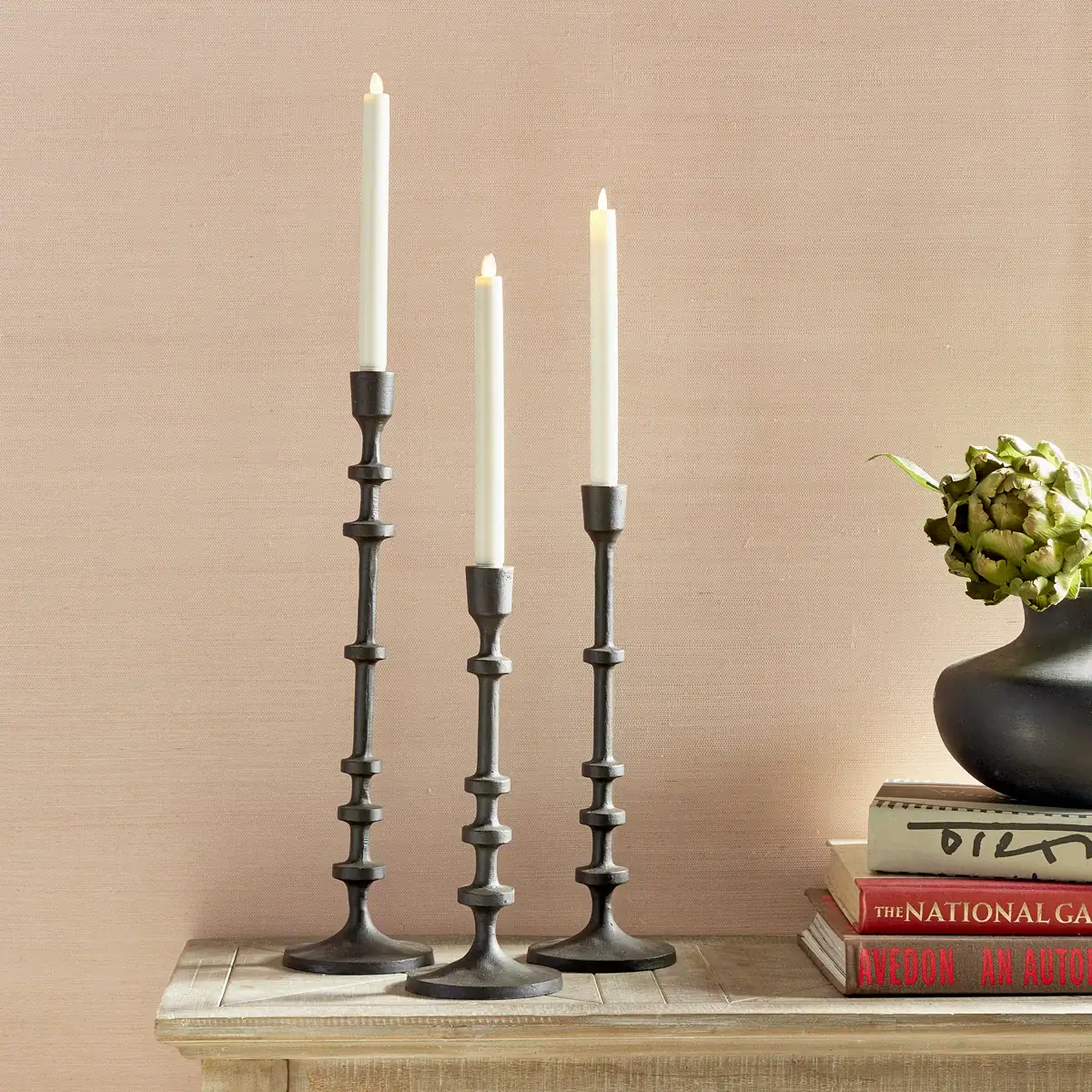 Three tall, black metal candlesticks with lit white taper candles stand on a wooden surface next to stacked books and a round vase holding a green artichoke, set against a pale pink wall.