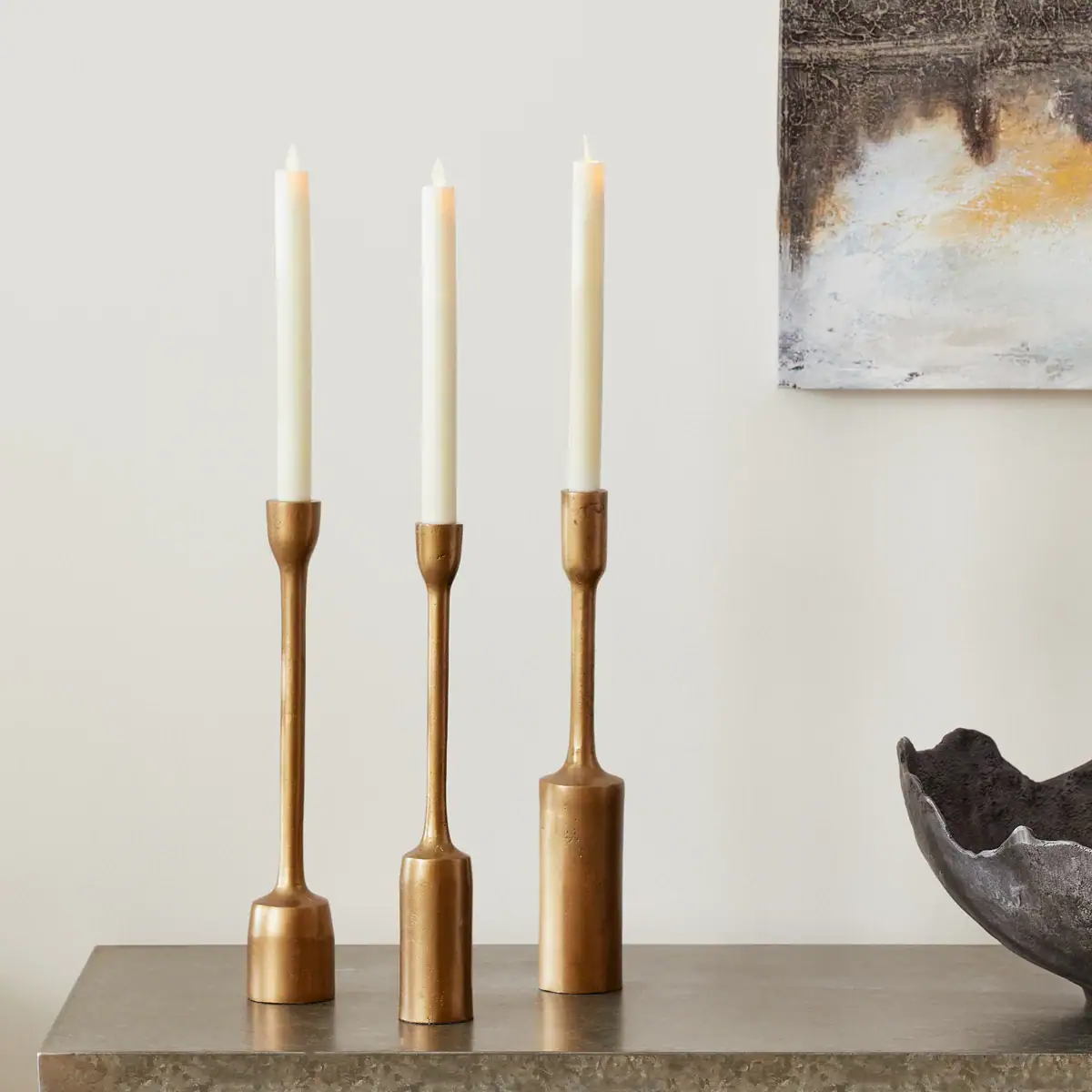 Three tall gold candle holders with white taper candles are arranged on a stone surface. In the background, part of an abstract painting and a dark decorative bowl are visible against a light-colored wall.