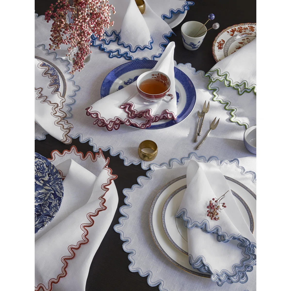 A table set with white napkins featuring colorful ruffled edges, patterned dishes, teacups, gold utensils, and small floral arrangements on a dark surface. The setting is elegant and decorative.