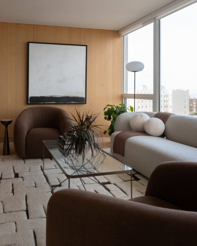A modern living room with wood-paneled walls, a large abstract painting, brown armchairs, a gray sofa with white pillows, a glass coffee table, some plants, and large windows with city views.